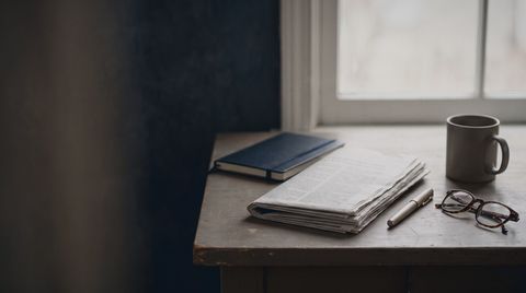 A desk by a window with a folded stack of papers, a pen and reading glasses
