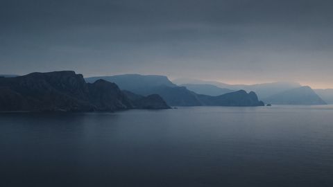 A rocky headland fading into blue haze over a calm, dark sea at dusk