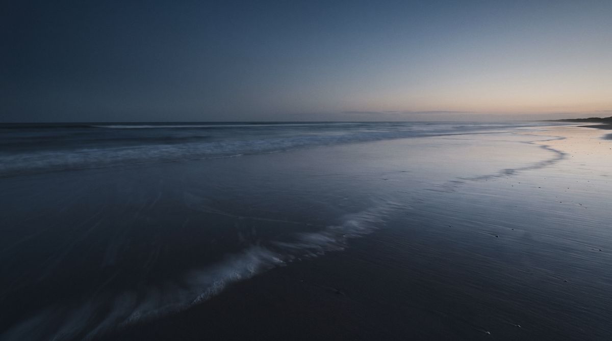 A dark, wet shoreline at blue hour with a thin band of light on the horizon